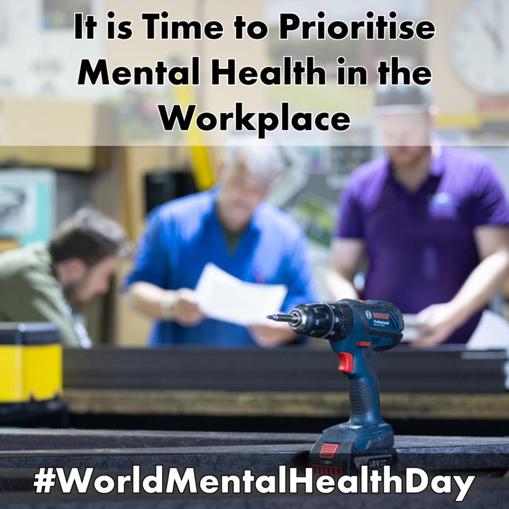 Slide 1 photo description: The Eco Furniture workshop with work benches. A power screwdriver is in focus in the foreground. Blurred in the background are 3 workers consulting design diagrams and wearing work clothes. Behind them is a blurred out notice board, clock, and boxes.
Slide 1 text: It is Time to Prioritize Mental Health in the Workplace  #WorldMentalHealthDay
